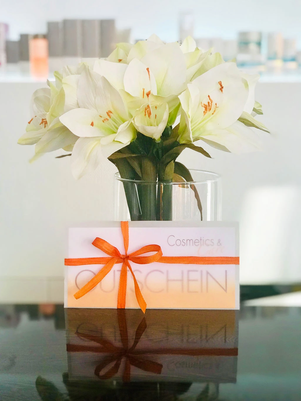 Ein Cosemtics & Care Gutschein verpackt in einen transparenten Umschlag mit einem Schleifchen steht angeleht an eine Blumenvase auf der Theke des Kosmetikstudios